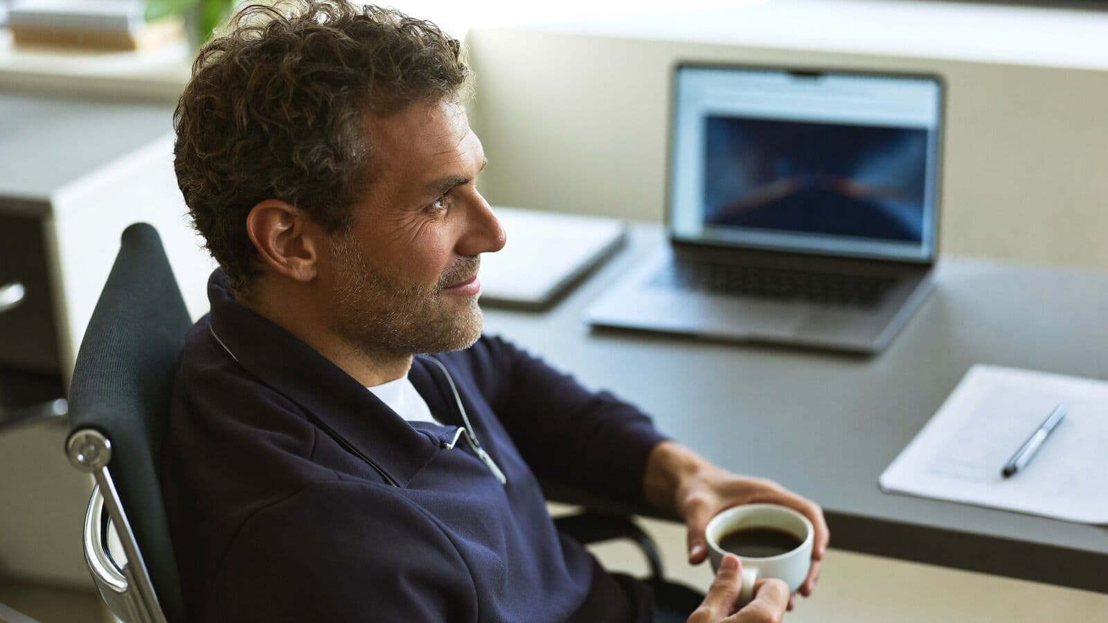 Business owner with a coffee looking out from his desk beside a laptop
