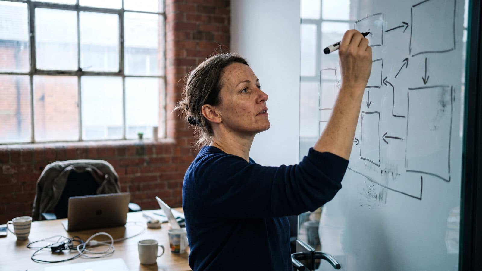 Business owner sketching sales pipeline stages on a glass wall beside a laptop showing an abstract kanban board